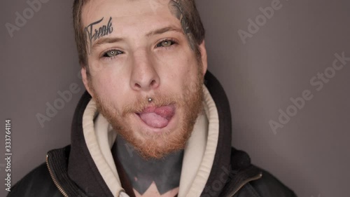Man with tattoo on his face and neck, eyeball or eye scleral tattooing, piercing demonstrating splitted tongue (body modification), posing in studio. Isolated on grey background. photo