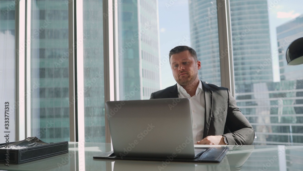 Young businessman in his office on the background of a skyscraper. Thoughtful young businessman in an elegant suit sitting on the background of a window in his office with skyscrapers Young businessman in his office on the background of a skyscraper. Thoughtful young businessman in an elegant suit sitting on the background of a window in his office with skyscrapers