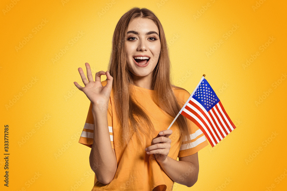 A beautiful young woman holds in her hands the flag of United States and shows the OK sign. Exchange student, learn English language. Tourist traveling. Football fan. A beautiful young woman holds in her hands the flag of United States and shows the OK sign. Exchange student, learn English language. Tourist traveling. Football fan.