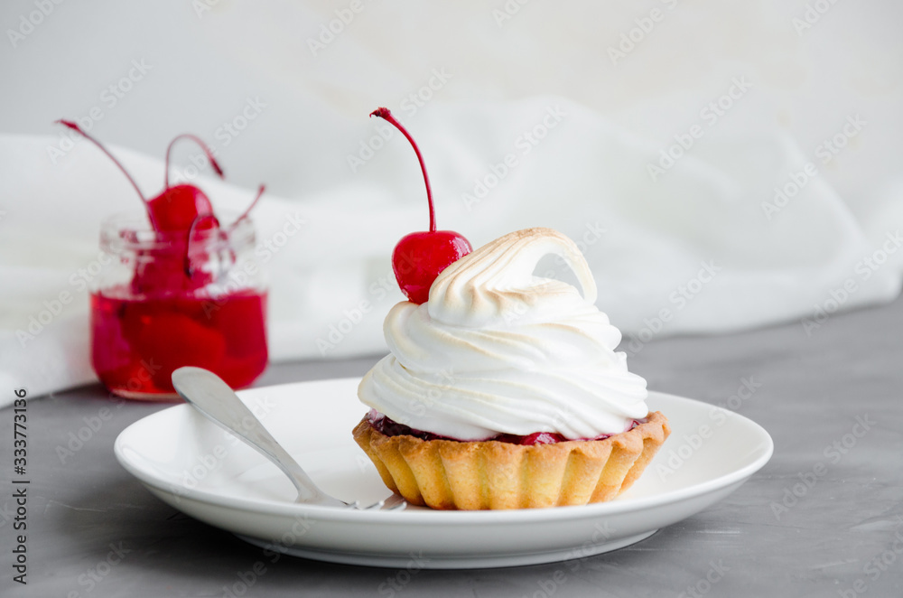 Tartalet with cherry filling and Italian meringue with a cocktail cherry on top on a white plate with a fork on a dark concrete background. Sweet food background. Cherry pie. Horizontal, Copy space. Tartalet with cherry filling and Italian meringue with a cocktail cherry on top on a white plate with a fork on a dark concrete background. Sweet food background. Cherry pie. Horizontal, Copy space.