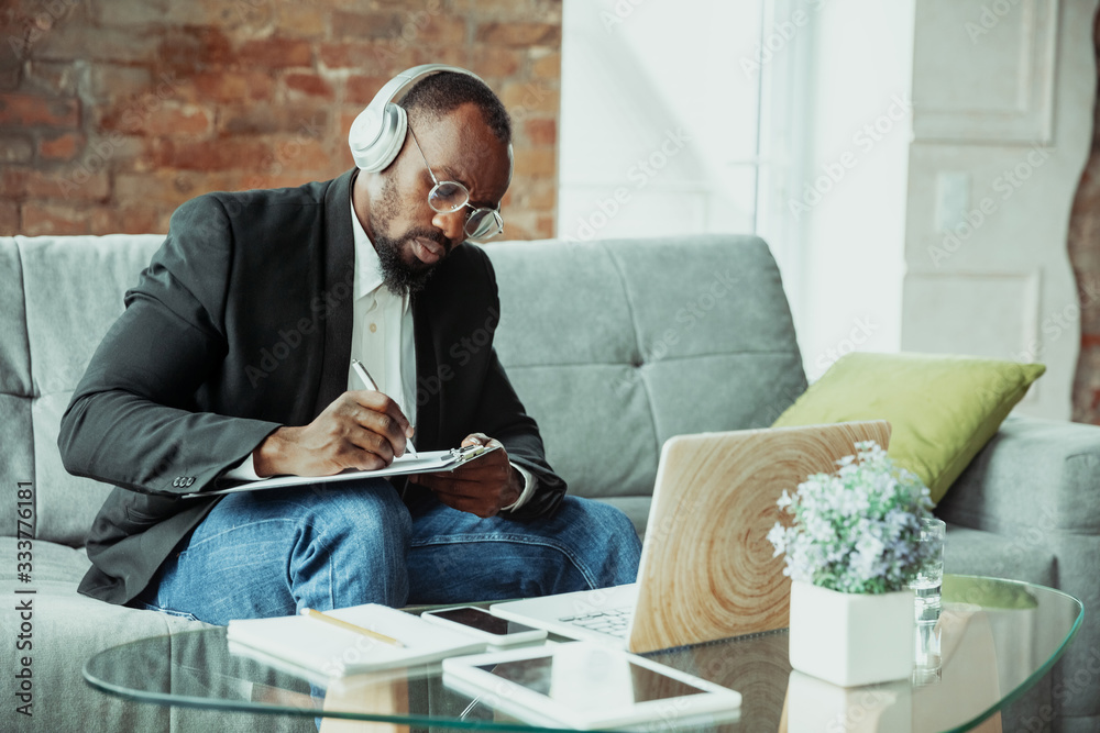 Businessman or student working from home being isolated or keep quarantine 'cause of coronavirus. African-american man using laptop, tablet and headphones. Online conference, lesson, remote office. - obrazy, fototapety, plakaty 