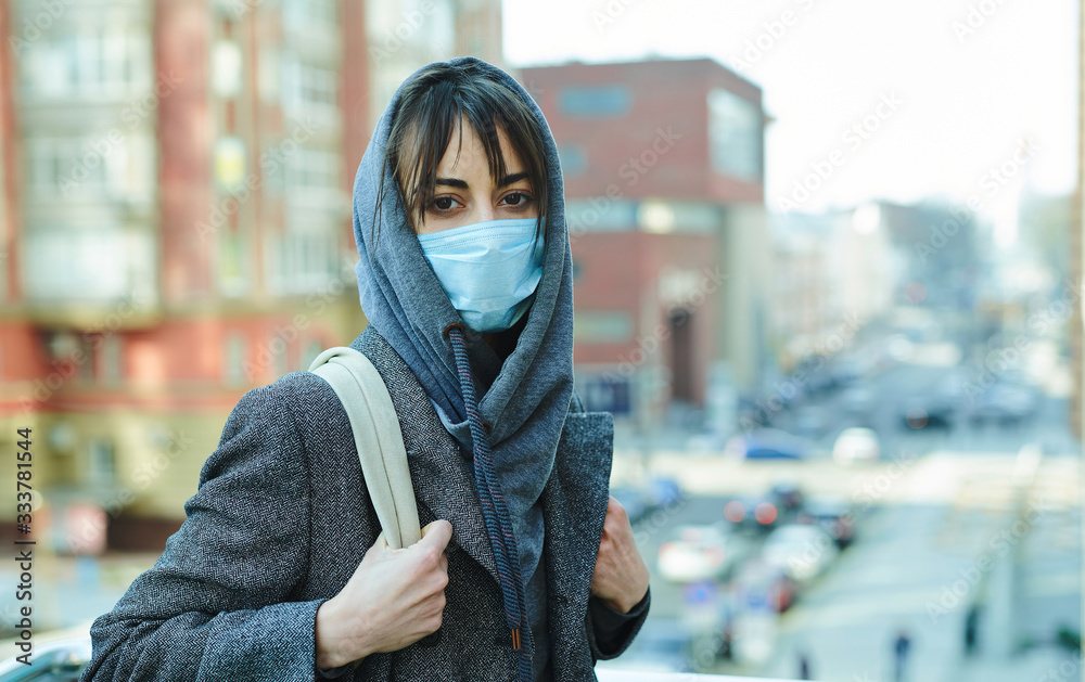 Woman in hood with backpack in city street with building on background, standing in protection face mask for prevention coronavirus. concept pandemic in city, COVID 19, quarantine, isolation period. Woman in hood with backpack in city street with building on background, standing in protection face mask for prevention coronavirus. concept pandemic in city, COVID 19, quarantine, isolation period.