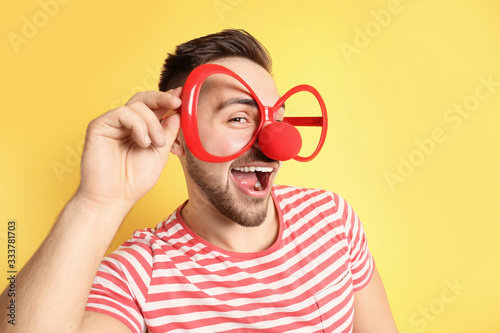 Emotional young man with party glasses and clown nose on yellow background. April fool's day