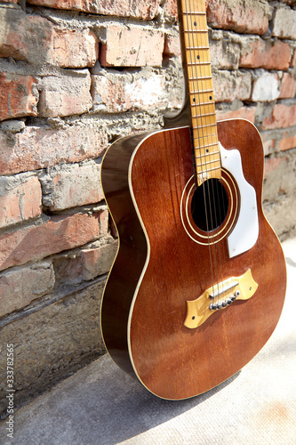 An old wooden guitar stands against a brick wall. The sun's rays fall on the guitar strings © Svetlana