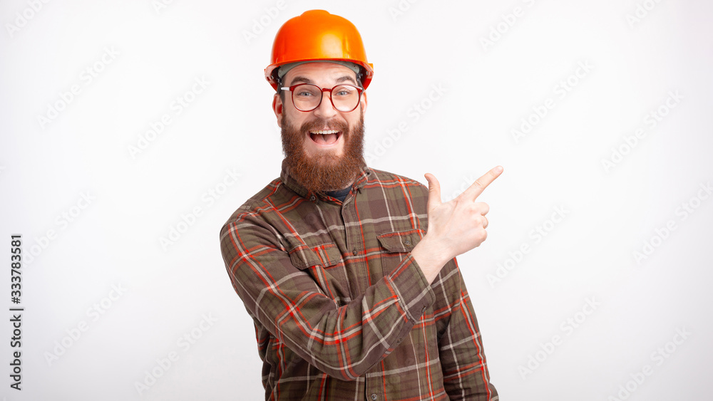 Young man wearing hard hat and glasses is pointing on his left on white background.