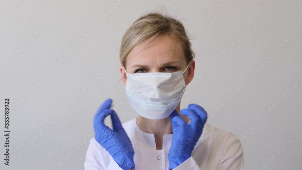 Blonde female doctor surgeon in white uniform and blue gloves puts on medical mask. Concept of hygiene, treatment, medicine. Blonde female doctor surgeon in white uniform and blue gloves puts on medical mask. Concept of hygiene, treatment, medicine.