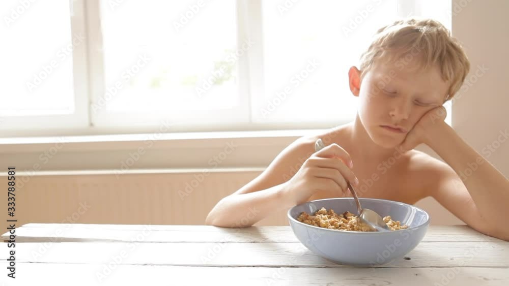 Sleepy boy sitting at table in morning before going to school idly stroking his porridge with spoon Sleepy boy sitting at table in morning before going to school idly stroking his porridge with spoon