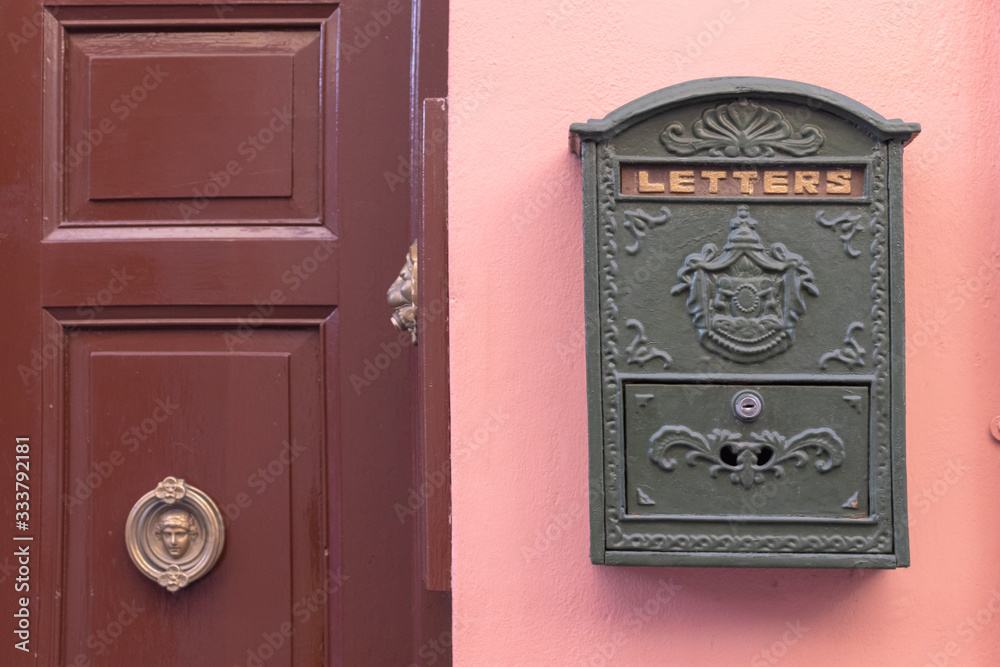 A bright retro looking green mailbox, or letterbox, affixed to the exterior wall of a pink house. There's a brown door with gold door knob A bright retro looking green mailbox, or letterbox, affixed to the exterior wall of a pink house. There's a brown door with gold door knob