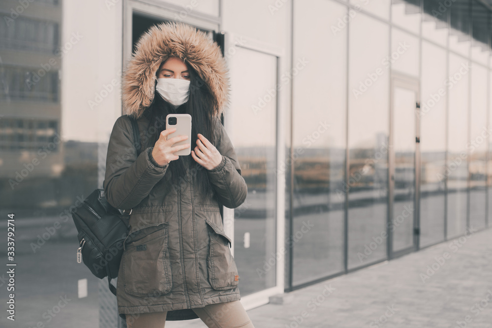 Fototapeta premium Woman using phone, wears a black protective medical face mask being in the city during a coronavirus pandemic. Protection from viruses in the city.