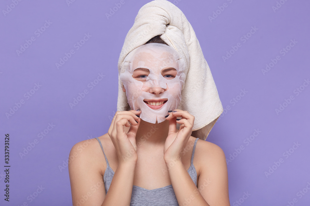 Horizontal shot of young adorable woman standing with face mask, posing isolated over lilac studio background, female looking at camera, taking care og her skin. Cosmetology and lifestyle concept. Horizontal shot of young adorable woman standing with face mask, posing isolated over lilac studio background, female looking at camera, taking care og her skin. Cosmetology and lifestyle concept.