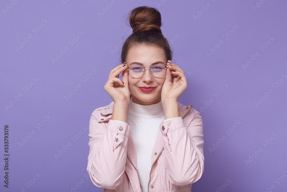 Studio portrait of woman with red lips, wearing stylish glasses, pink jacket and white, playfully looking at camera and tounching frame of her glasses, standing isolated over lilac studio background. Studio portrait of woman with red lips, wearing stylish glasses, pink jacket and white, playfully looking at camera and tounching frame of her glasses, standing isolated over lilac studio background.