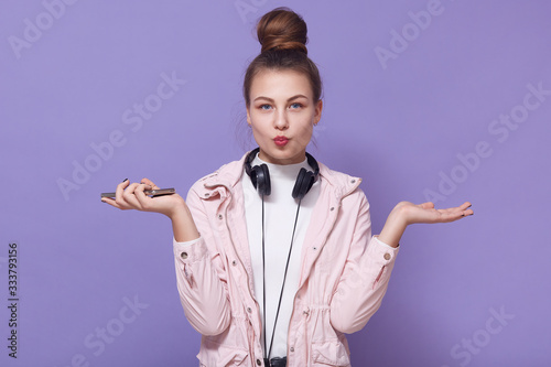 Indoor shot of teenager caucasian woman wears white shirt and pink jacket, posing isolated over lilac wall, looks directly atcamera, makes kiss gesture, spreads palms aside, has headphone around neck. photo