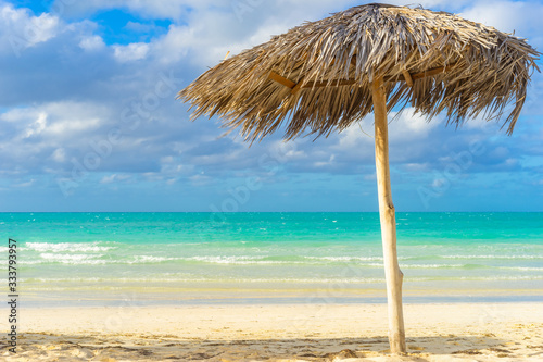 Caribbean landscape with beach umbrella. Vacation in Cuba. Palm leaf umbrella on the Cuban beach. Rest from all worries. Paradise. Travel to Cuba. White sand and emerald water of the Caribbean sea.
