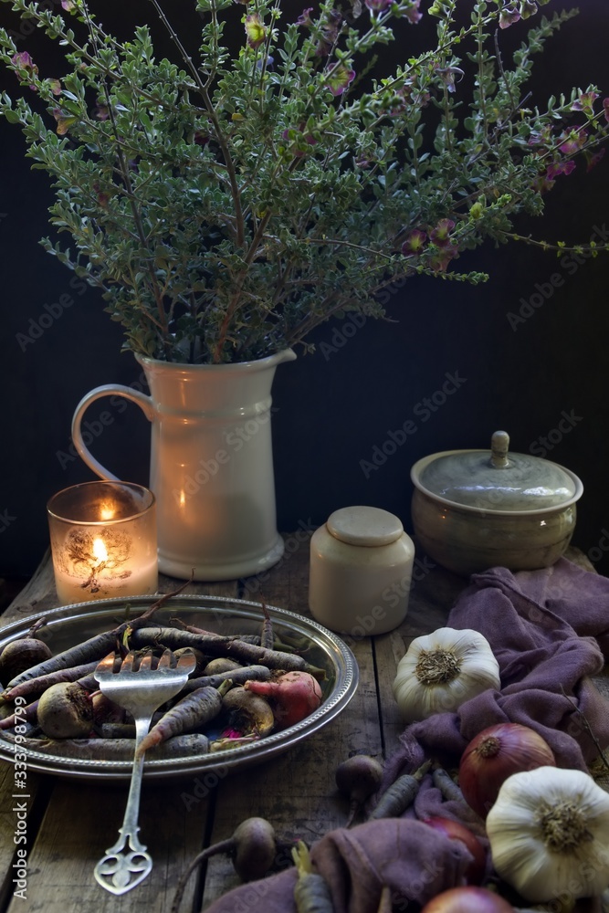 Still life seen of root vegetables in a silver platter with a silver serving fork.  Jug of flowers and pots with a lit candle sit in the background.  Garlic bulbs and red onions sit to the right.