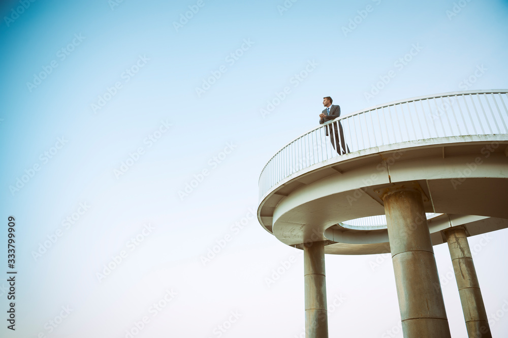 Distant businessman standing in isolation on a curved elevated walkway under soft blue sky 