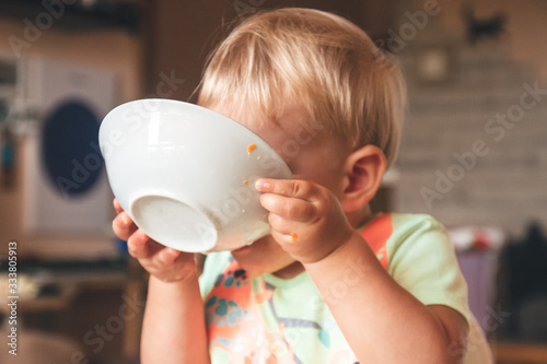 blond kid boy, very hungry eats and holds a plate with both hands, is covered with a plate, close-up