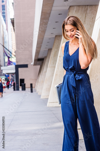 Obraz na płótnie Young Eastern European American Woman talking on cell phone, traveling, working in New York City, wearing blue jumpsuit, carrying laptop computer, looking down, walking out from office building.. w salonie