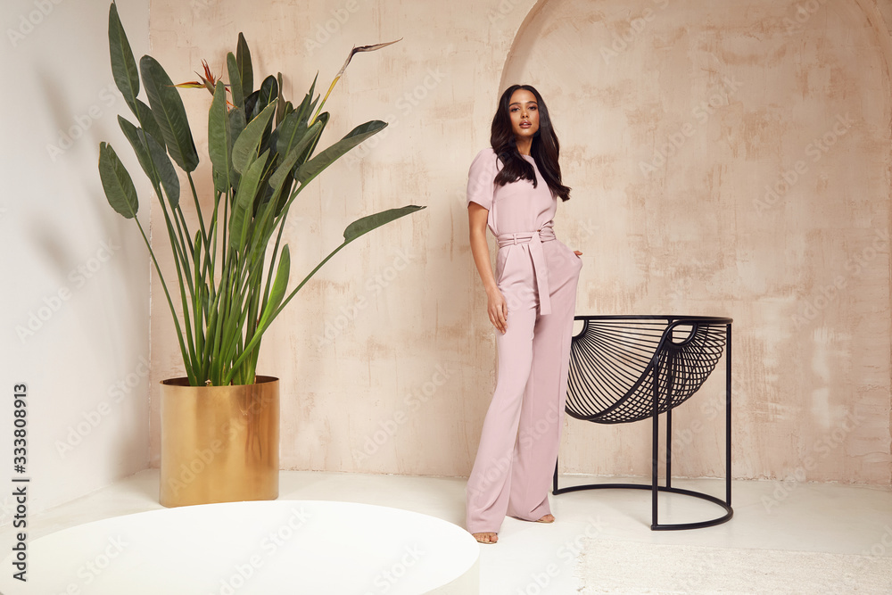 Beautiful sexy brunette woman tanned skin face cosmetic makeup wear pink suit pants for date walk office fashion clothes style collection interior room  sand color safari summer armchair palm boho. - obrazy, fototapety, plakaty 