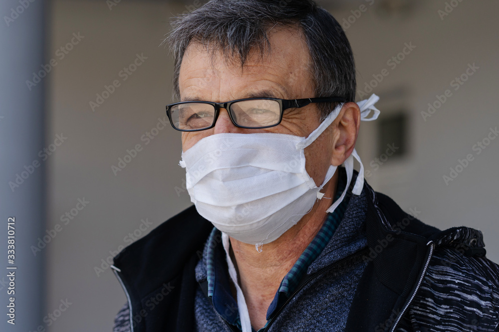Front view portrait caucasian senior man standing outdoor in front of the building or home wearing medical protective mask to prevent pandemic disease spread protecting from viruses pollution in day