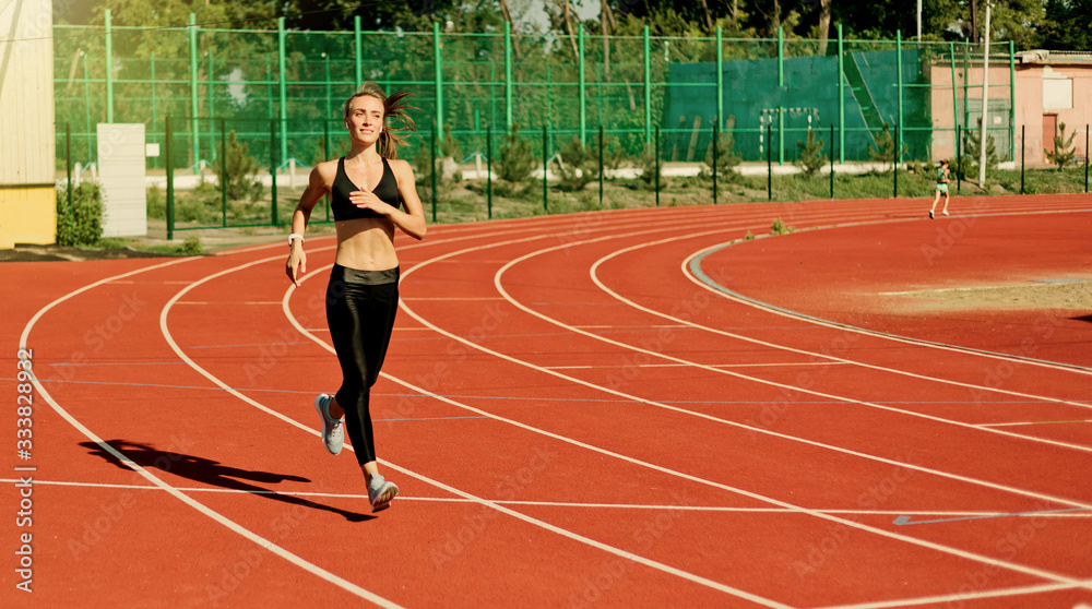 Young cheerful woman runner in sportswear running on stadium track with red coating outdoors