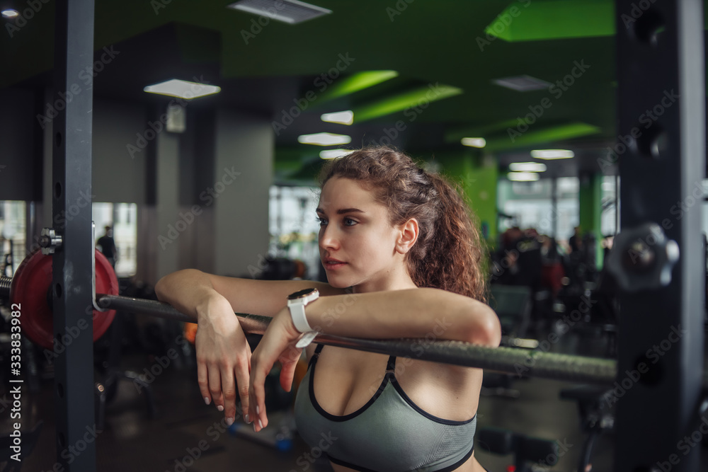 Fototapeta premium Tired fit woman in sportswear leaning on barbell in the gym