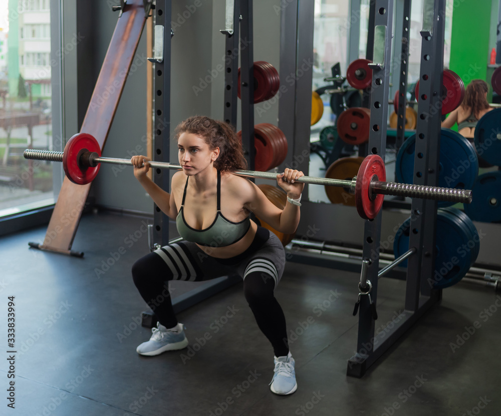 Fototapeta premium Young fit woman in sportswear crouches with a barbell on her shoulders in the gym. Healthy lifestyle concept. Hard training