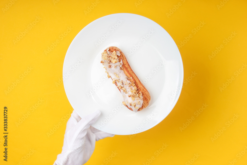 Waiter in white gloves serves eclair on a plate. Yellow background. Dessert. Restaurant food. Top view, minimalism