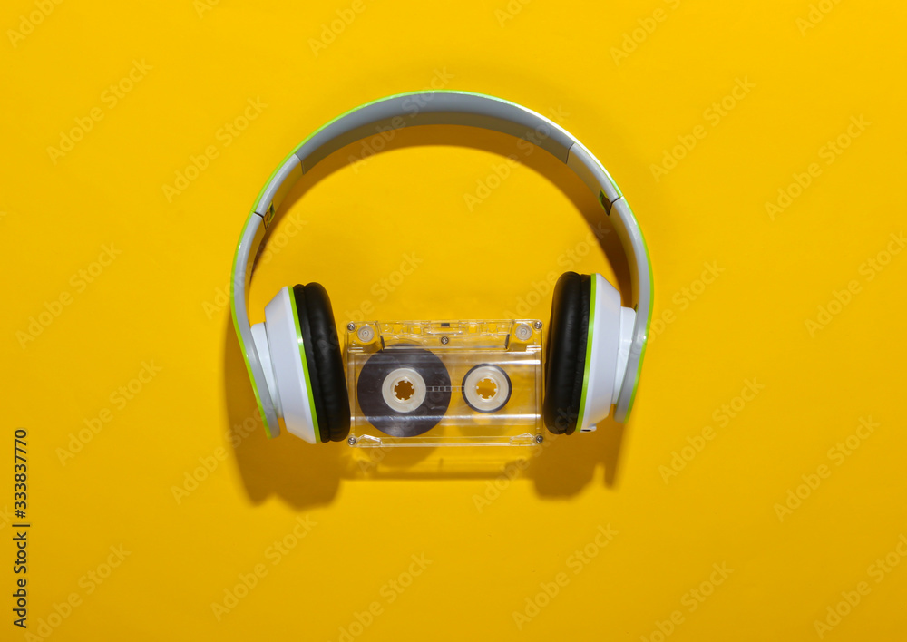 Stylish wireless stereo headphones and audio cassette on yellow background with shadow. Music lover. Retro 80s. Top view. - obrazy, fototapety, plakaty 