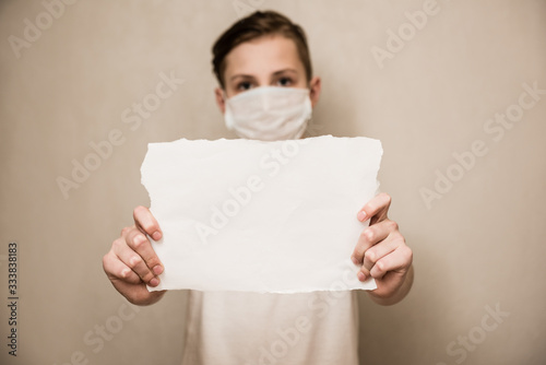 Portrait of caucasian boy in a protective mask respirator holds a blank sheet of paper on gray background. Protection against coronavirus. Mock up, copy space, advertisement photo