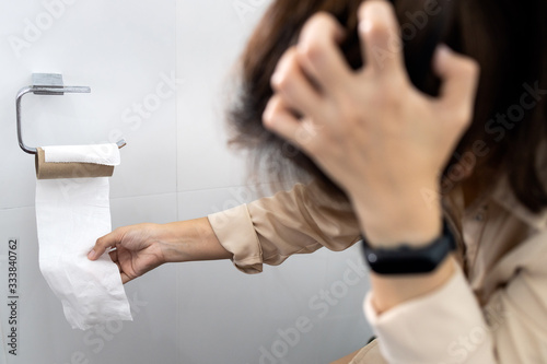 Stressed woman sitting on toilet bowl with tissue roll that was about to run out effects from the Coronavirus problem of tissue paper is out of stock during outbreak of Covid-19 out of toilet paper