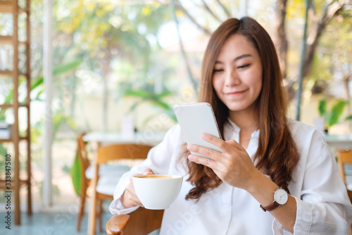 Closeup image of a beautiful asian woman holding and using mobile phone while drinking coffee in cafe