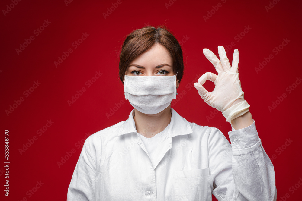 Young beautiful caucasian female in blue medical gown and with white medical mask on her face looks on the camera and shows OK