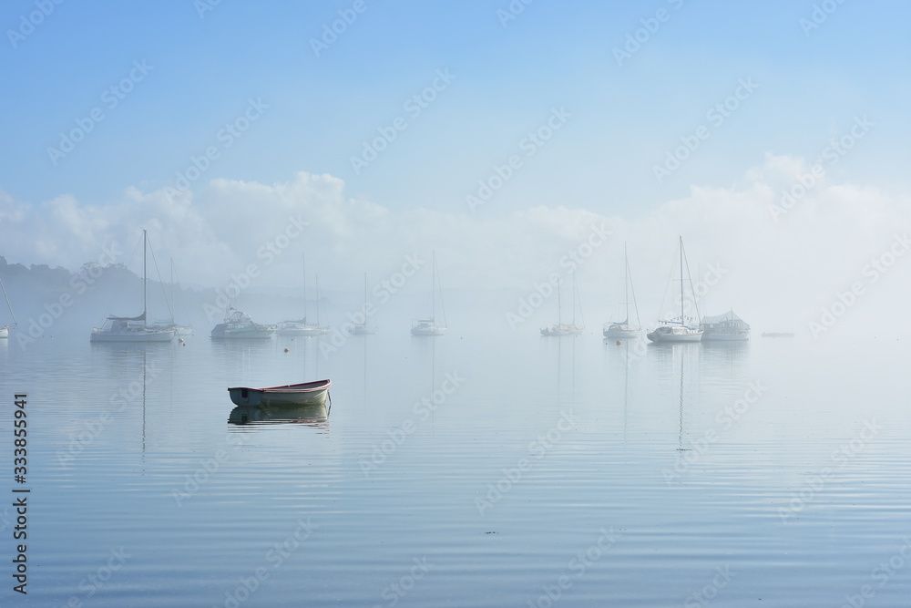 Obraz premium Lonely dinghy anchored on flat calm water with boats moored in background in morning fog.