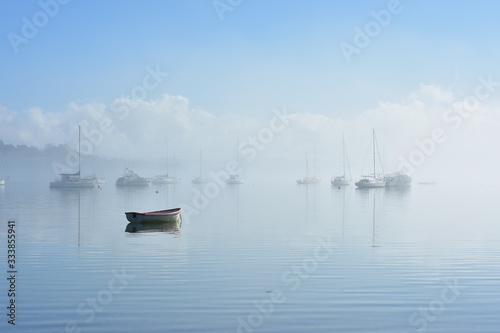 Lonely dinghy anchored on flat calm water with boats moored in background in morning fog.