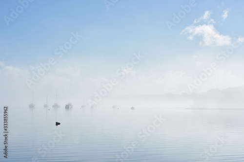 Lonely buoy on flat calm water marking anchorage with boats moored in background in morning fog.
