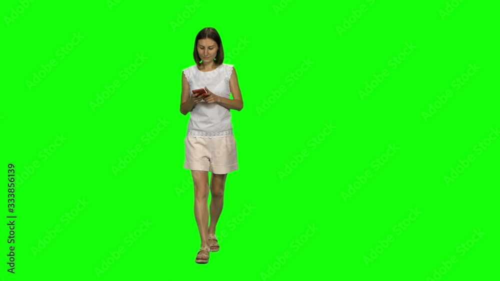 Young woman is calmly walking and texting message vie her mobile phone on green screen. Front view.