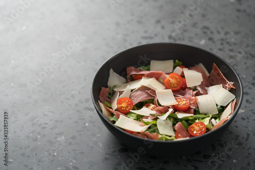 Salad with prosciutto, tomato, arugula leaves and parmesan cheese in black bowl on concrete surface