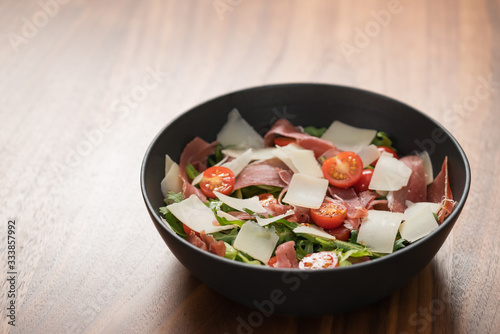 Salad with prosciutto, tomato, arugula leaves and parmesan cheese in black bowl on walnut