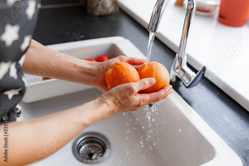 Hands of woman washing ripe orange under faucet in the sink kitchen