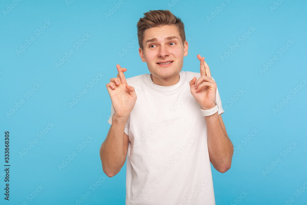 In anticipation of win. Portrait of man in casual white t-shirt crossing fingers in good luck gesture and looking with hope, making wish to success. indoor studio shot isolated on blue background In anticipation of win. Portrait of man in casual white t-shirt crossing fingers in good luck gesture and looking with hope, making wish to success. indoor studio shot isolated on blue background