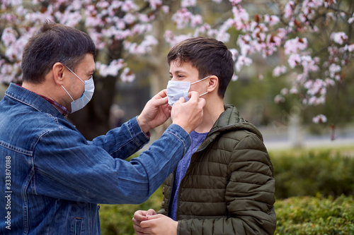father and son with a face mask are in the city outdoor, blooming trees, spring season, flowering time - concept of allergies and health protection from dusty air