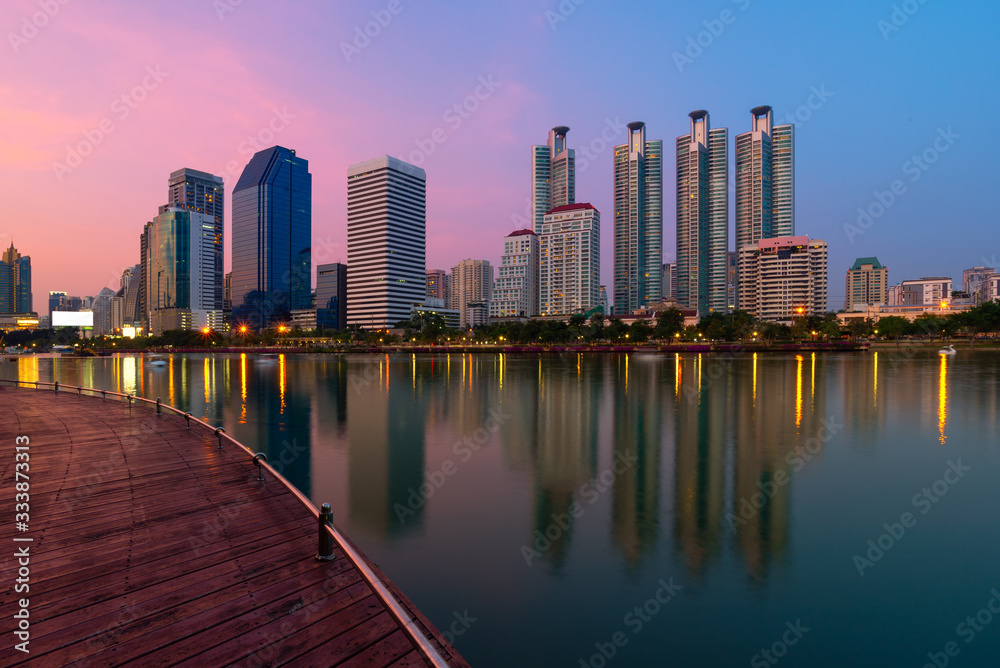 Naklejka premium Landscape or cityscape ofpublic park with wooden walk way path, lake and urban building reflection with water surface at twilight time.