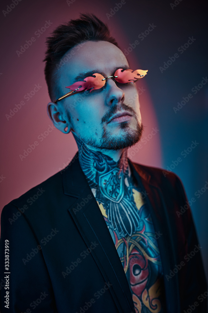 Extravagant tattoo artist posing in a neon studio wearing a black tuxedo on a half-naked body, fire-shaped sunglasses and tattooed in a japanese irezumi style, looking self-assured. Vertical portrait Extravagant tattoo artist posing in a neon studio wearing a black tuxedo on a half-naked body, fire-shaped sunglasses and tattooed in a japanese irezumi style, looking self-assured. Vertical portrait