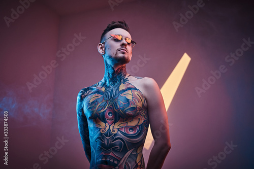 Expressive male model posing in a neon studio with a half-naked body wearing fire-shaped sunglasses and tattooed in a japanese irezumi style, looking cool and relaxed.