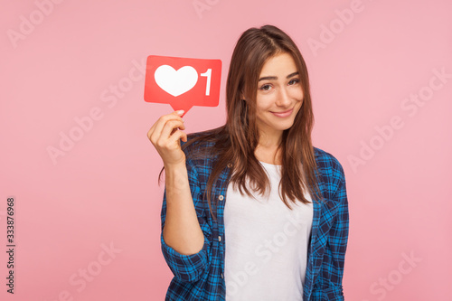Portrait of cute positive girl in checkered shirt smiling playfully and holding social media like icon, heart button to enjoy trendy interesting content. indoor studio shot isolated on pink background