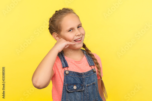 Portrait of funny positive little girl with braid in denim overalls making call with fingers shaped like telephone near head, communicating by phone. indoor studio shot isolated on yellow background