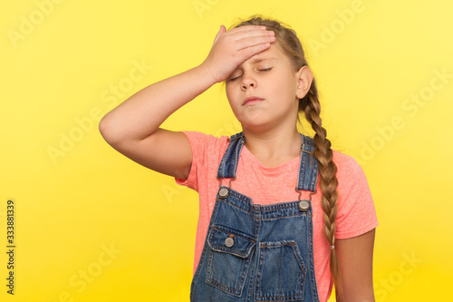 Fotografi Portrait of forgetful sad little girl with braid in denim overalls gesturing facepalm with hand on forehead and expressing sorrow, blaming herself for bad memory
