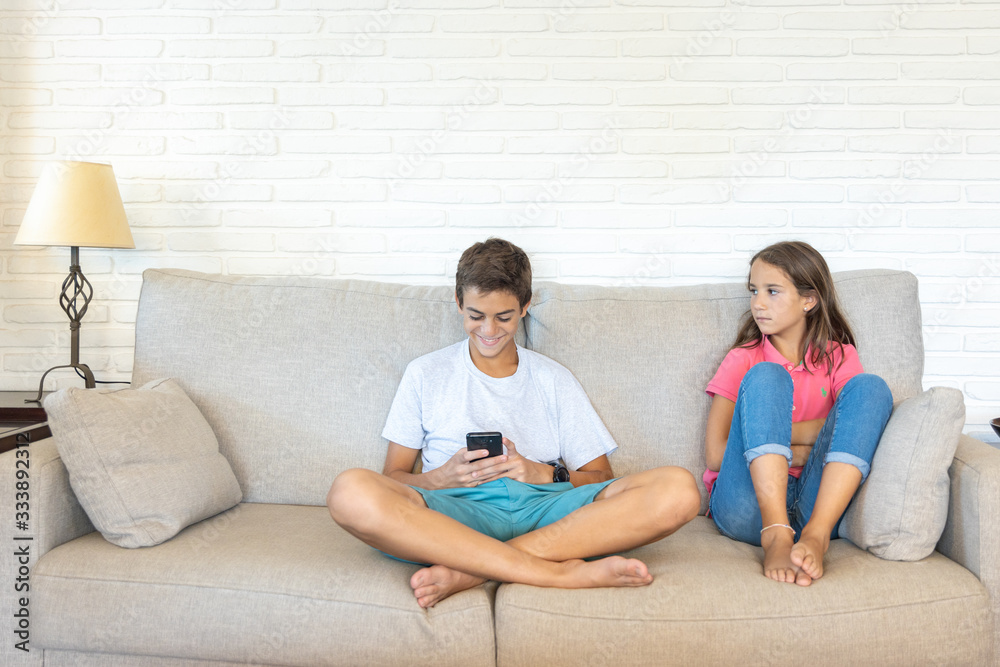 Happy boy playing with smartphone next to his bored sister looking at him Happy boy playing with smartphone next to his bored sister looking at him