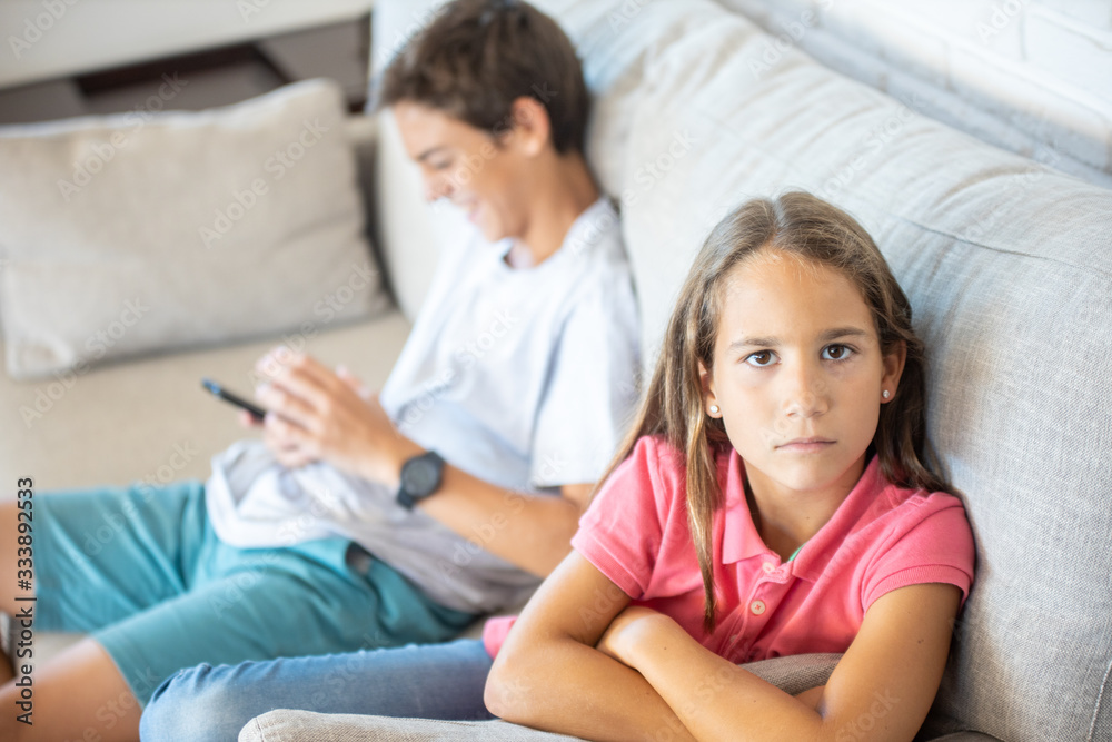 Happy boy playing on smartphone next to his angry sister looking at camera Happy boy playing on smartphone next to his angry sister looking at camera