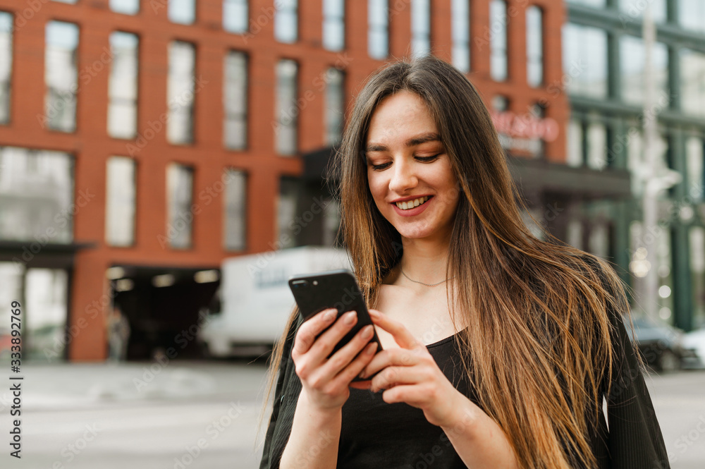 Happy lady in formal wear uses a smartphone and looks at a smartphone screen with a smile on her face. Cute smiling businesswoman communicates in a smart phone while walking through the city.